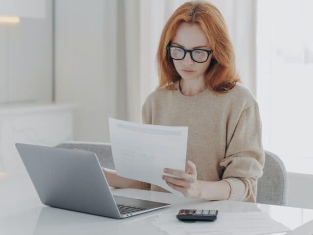 Young focused red-haired woman sitting at table with laptop and holding paper taxes bills